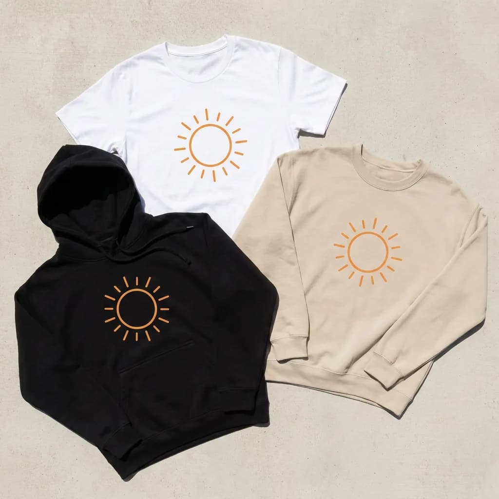 A merch collection flat lay of a white t-shirt, a black hoodie and a sand-colored sweatshirt, all printed with the same minimal orange sun logo, on a light concrete background