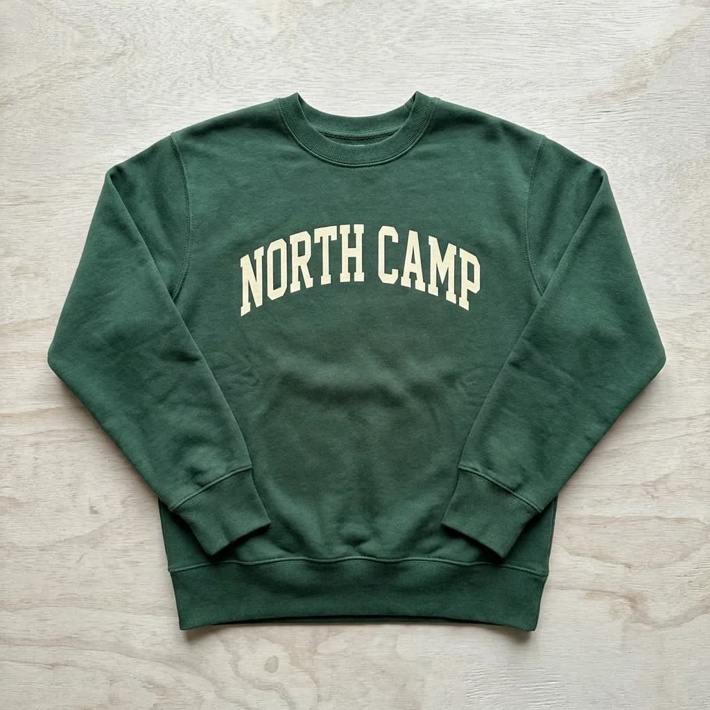 A realistic forest green crewneck sweatshirt mockup lying flat on a light background with a cream varsity-style arched 'NORTH CAMP' graphic printed on the chest and visible fabric texture